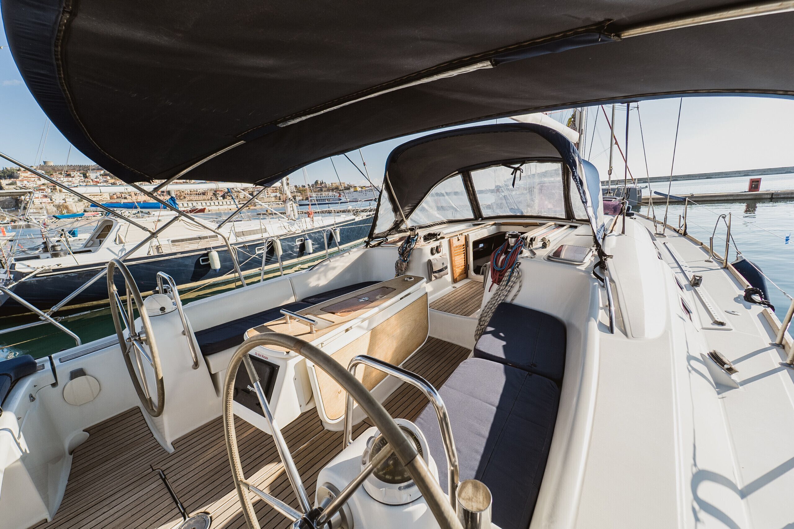 A sailboat's deck with a steering wheel, a cushioned seating area, and a view of the marina.