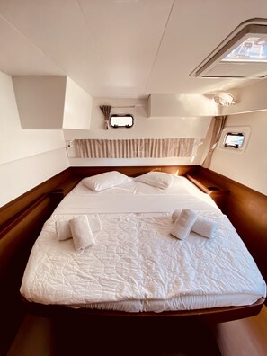A bed with white linens, two pillows, and two towels. There are two windows with curtains, a headboard, and a small shelf with a lamp. - Sun-Kissed Sailing Adventure in the Ionian Islands, Lefkada (Lefkada)