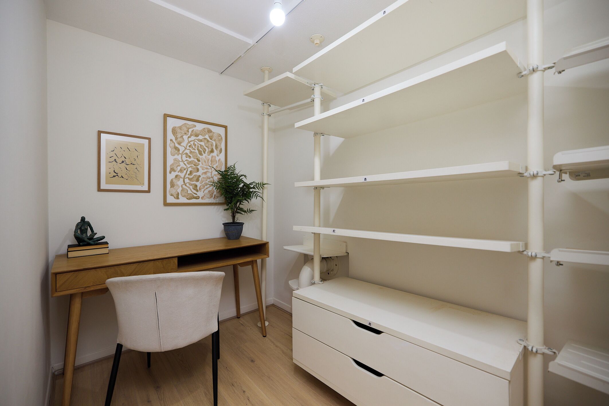 Una habitación con un escritorio de madera, una silla blanca, un estante blanco y dos cuadros en la pared.