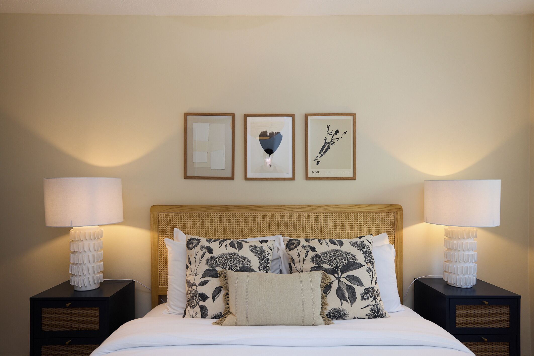 Una cama bien tendida con almohadillas estampadas con motivos florales, un cabecero y dos mesitas de noche con lámparas. Tres cuadros enmarcados cuelgan de la pared sobre la cama.