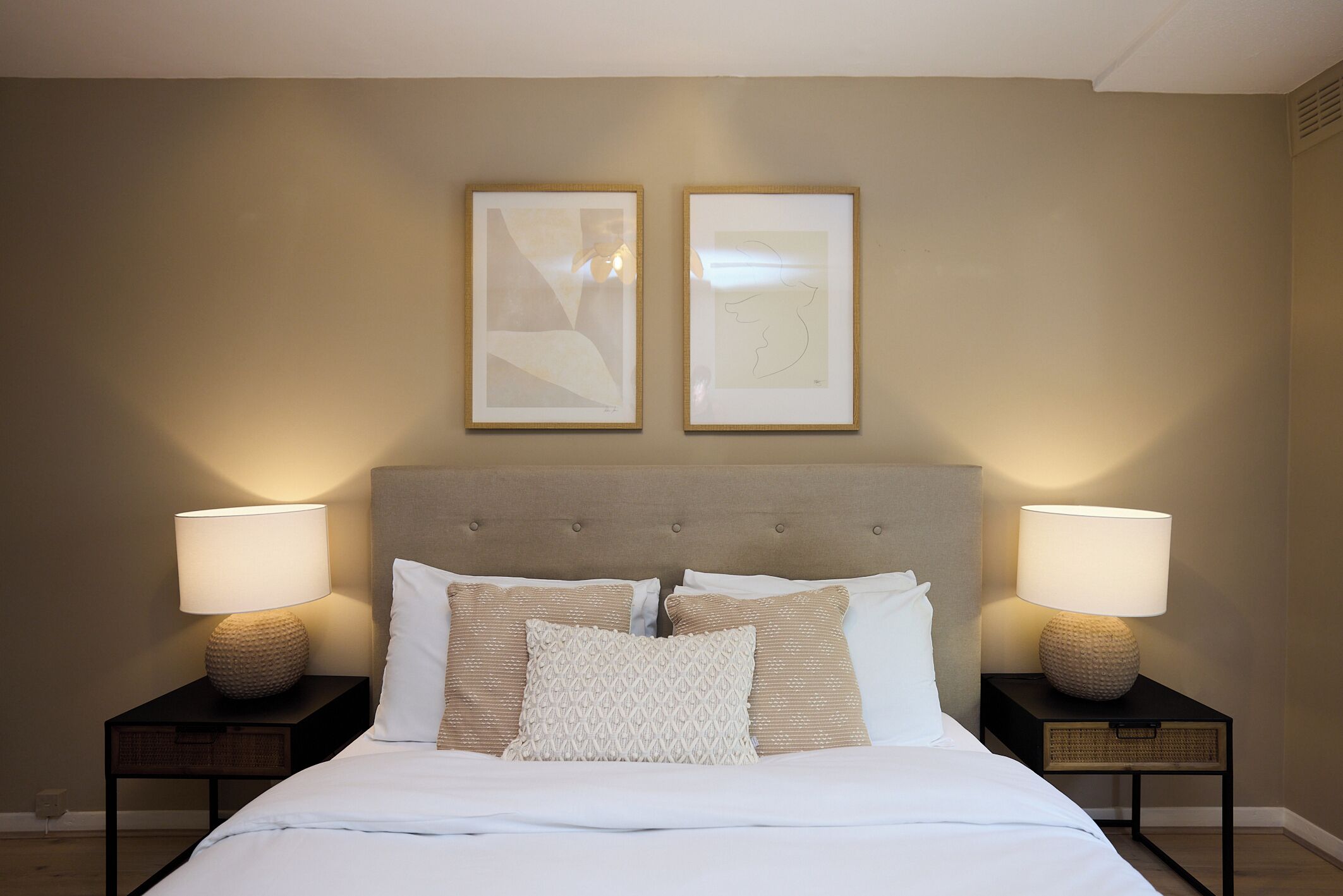 A neatly made bed with two bedside lamps, a headboard, and two framed artworks on the wall.