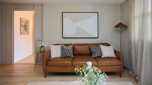 A living room with a brown leather sofa, a framed abstract painting, a floor lamp, and a small side table with a vase. - The Westbourne Side Story (London)
