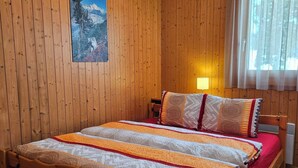 A wooden-paneled bedroom with a bed, a nightstand, a lamp, and a window with curtains. - Amiel EG Süd Appartement/Fewo, Dusche und Bad, WC 3 1/2 Zimmerwohnung, 5-Betten by Interhome (Riederalp)