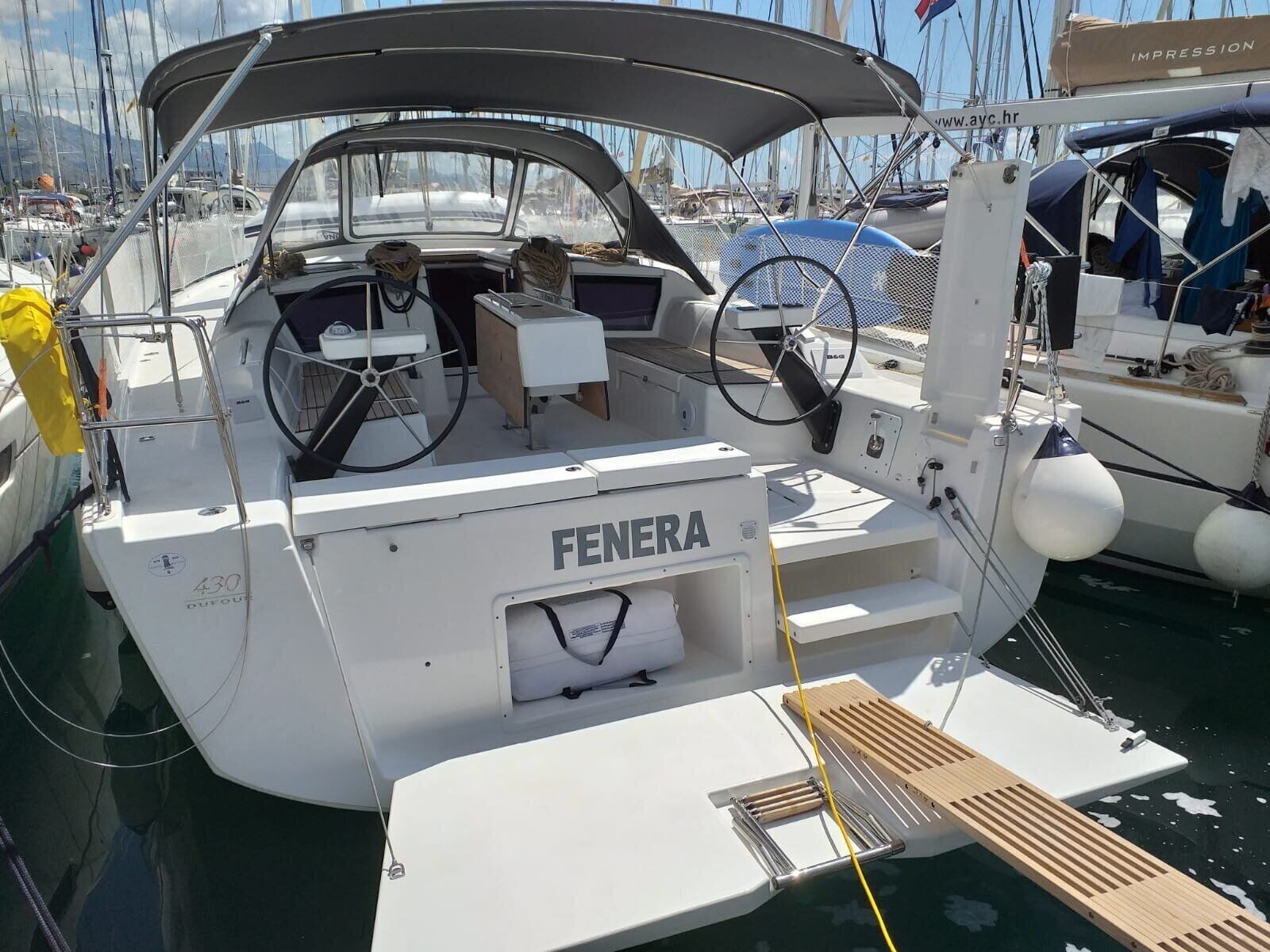 A white boat named 'FENERA' docked at a marina with a covered cockpit, steering wheel, and a small table.