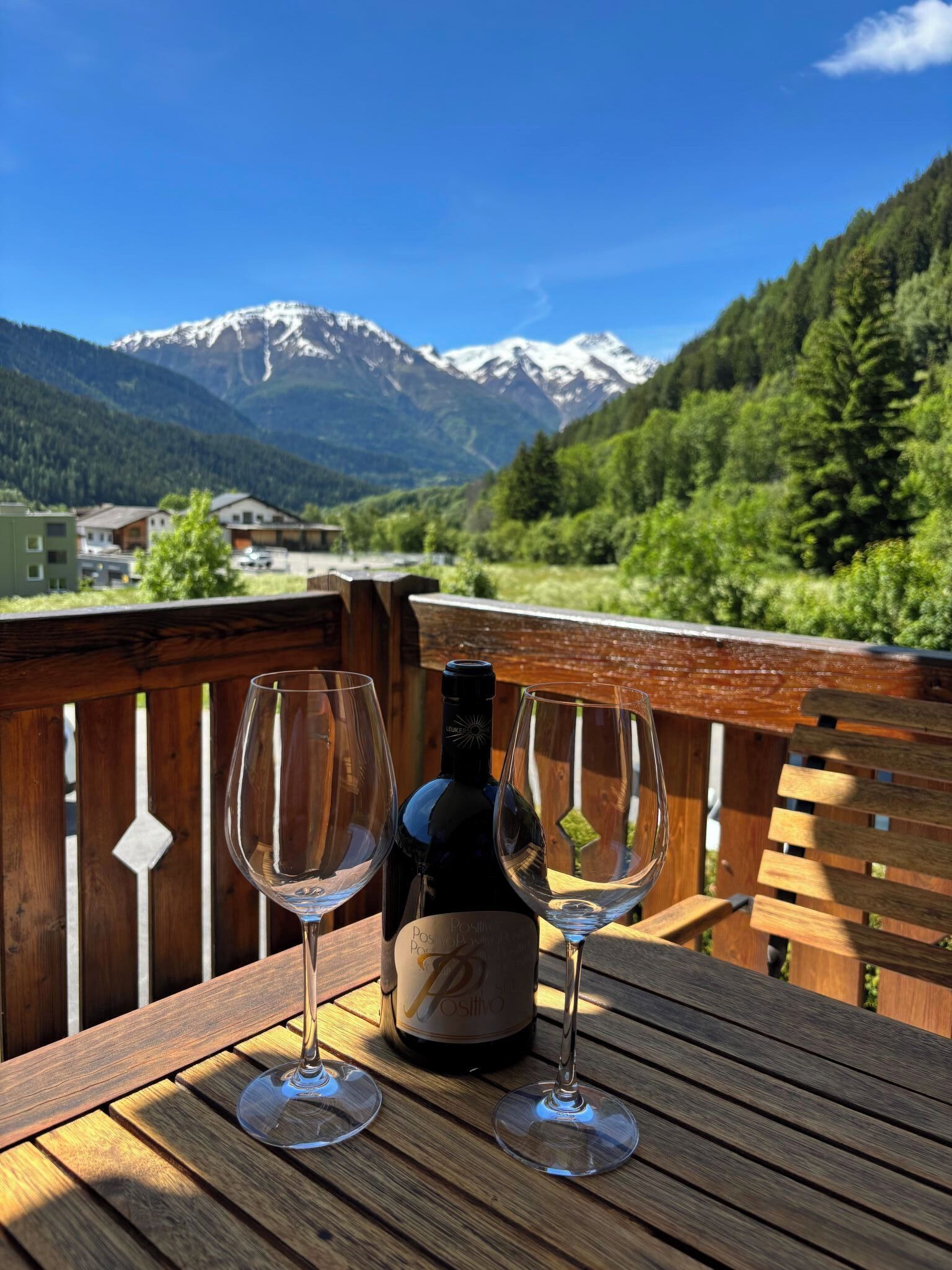 A wooden table with two wine glasses and a bottle of wine, set against a backdrop of a mountain landscape.