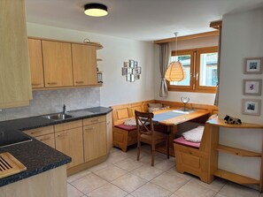 A compact kitchen with wooden cabinets, a built-in bench with a cushioned seat, a sink, and a window with curtains.