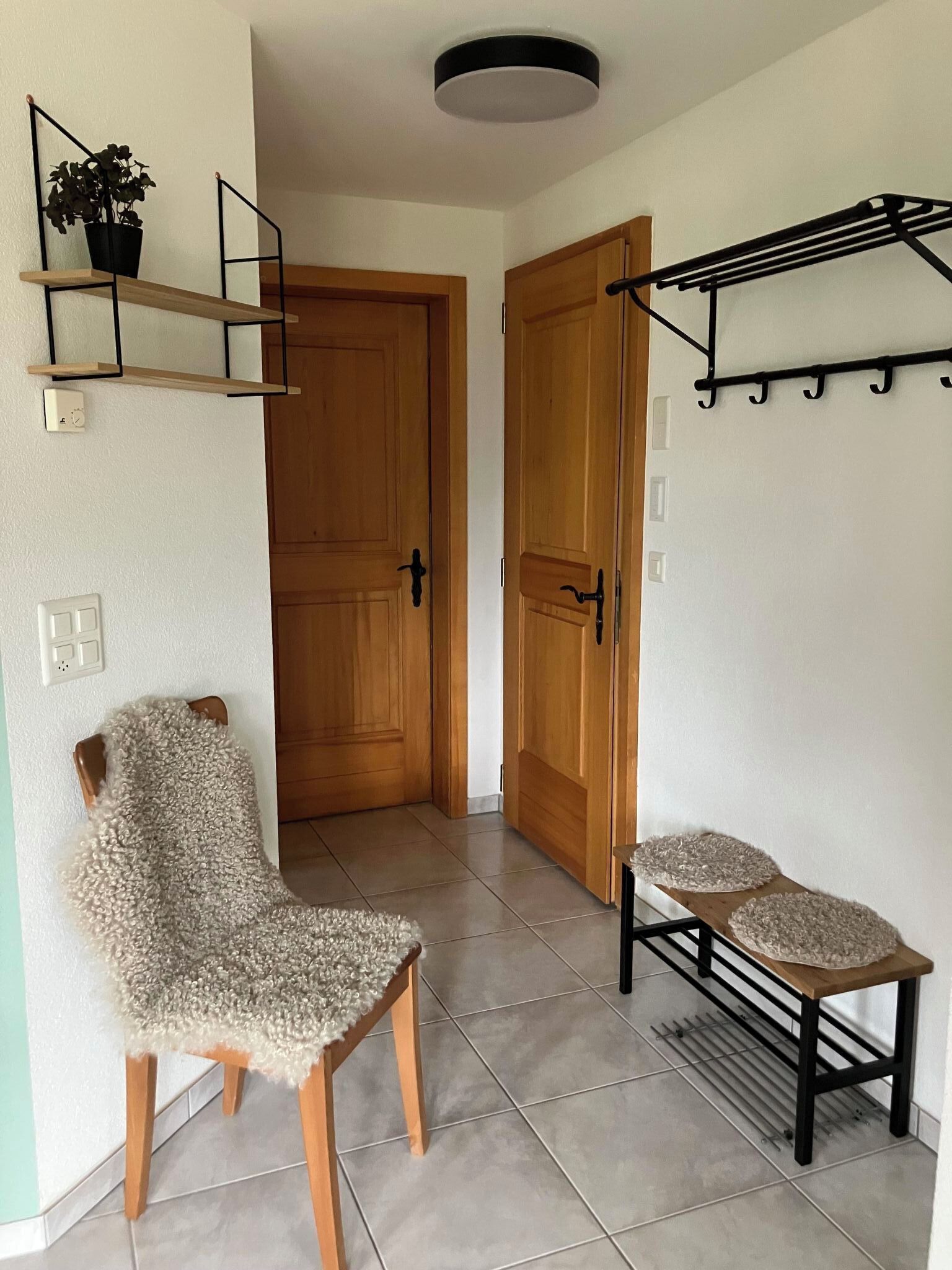 Un couloir avec une porte en bois, une chaise avec un siège texturé, une petite table recouverte d’une nappe décorative et un étendoir fixé au mur.
