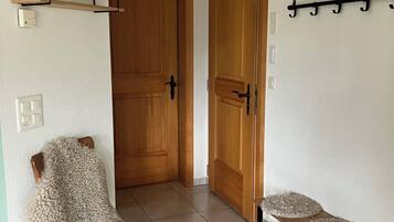 Un couloir avec une porte en bois, une chaise avec un siège texturé, une petite table recouverte d’une nappe décorative et un étendoir fixé au mur.