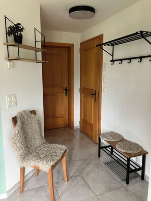 A hallway with a wooden door, a chair with a textured seat, a small table with a decorative cover, and a wall-mounted drying rack.