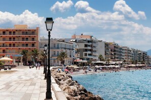 Eine Küstenpromenade mit Steinweg, einem Strand mit Sonnenschirmen und mehrstöckigen Gebäuden im Hintergrund.