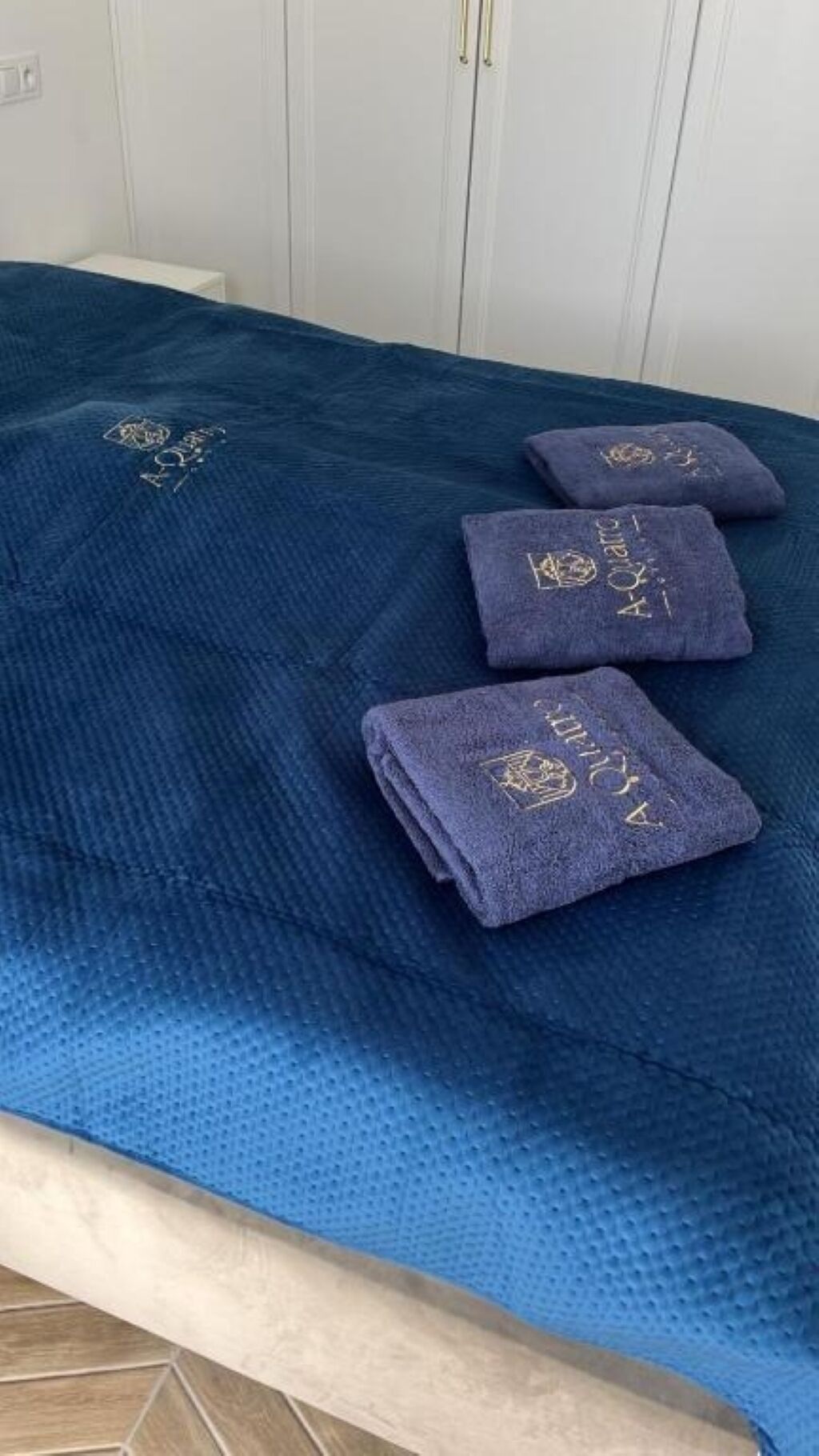 A bed with a blue textured bedspread and three folded towels with embroidered text.