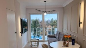 A modern living room with a sofa, round dining table, chairs, a TV mounted on the wall, and a balcony with a railing.