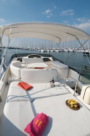 A boat with a white cushioned seating area, a table with a bottle and glasses, and a basket of fruit. - Sun-Kissed Sailing Adventure in Islas Sarónicas (Alimos)