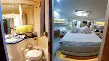 A compact, well-lit bathroom with a sink, toilet, and towel rack. The room features a bed with a headboard and pillows, a bedside table, and a mirror.