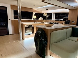 A modern kitchen with wooden cabinetry, a built-in oven, a sink, and a dining area with a light-colored sofa. - Sun-Kissed Sailing Adventure in Islas Sarónicas (Alimos)