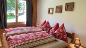 Una cama de madera con ropa de cama estampada y almohadas con forma de corazón, una mesita de noche con lámpara y una ventana con cortinas.