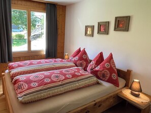 Ein Bett aus Holz mit gemusterten Textilien und herzförmigen Kissen, ein Nachttisch mit Lampe und ein Fenster mit Vorhängen.