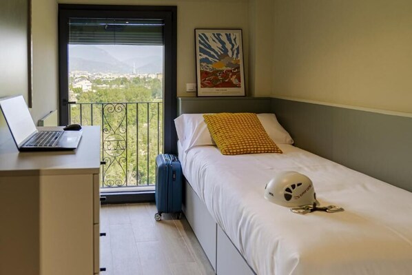 Habitación de hotel con cama, escritorio con laptop, maleta, casco sobre la cama y balcón con vistas.