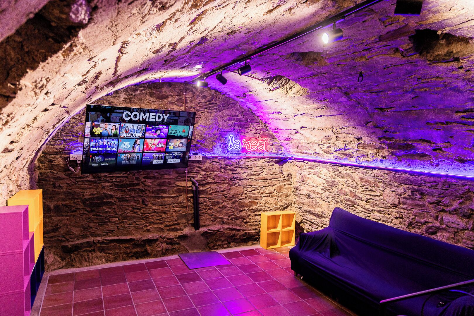 Una habitación acogedora con pared de piedra, un televisor de pantalla plana mostrando un canal de comedia, un sofá morado y un pequeño estante de madera.