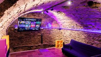 Una habitación acogedora con pared de piedra, un televisor de pantalla plana mostrando un canal de comedia, un sofá morado y un pequeño estante de madera.