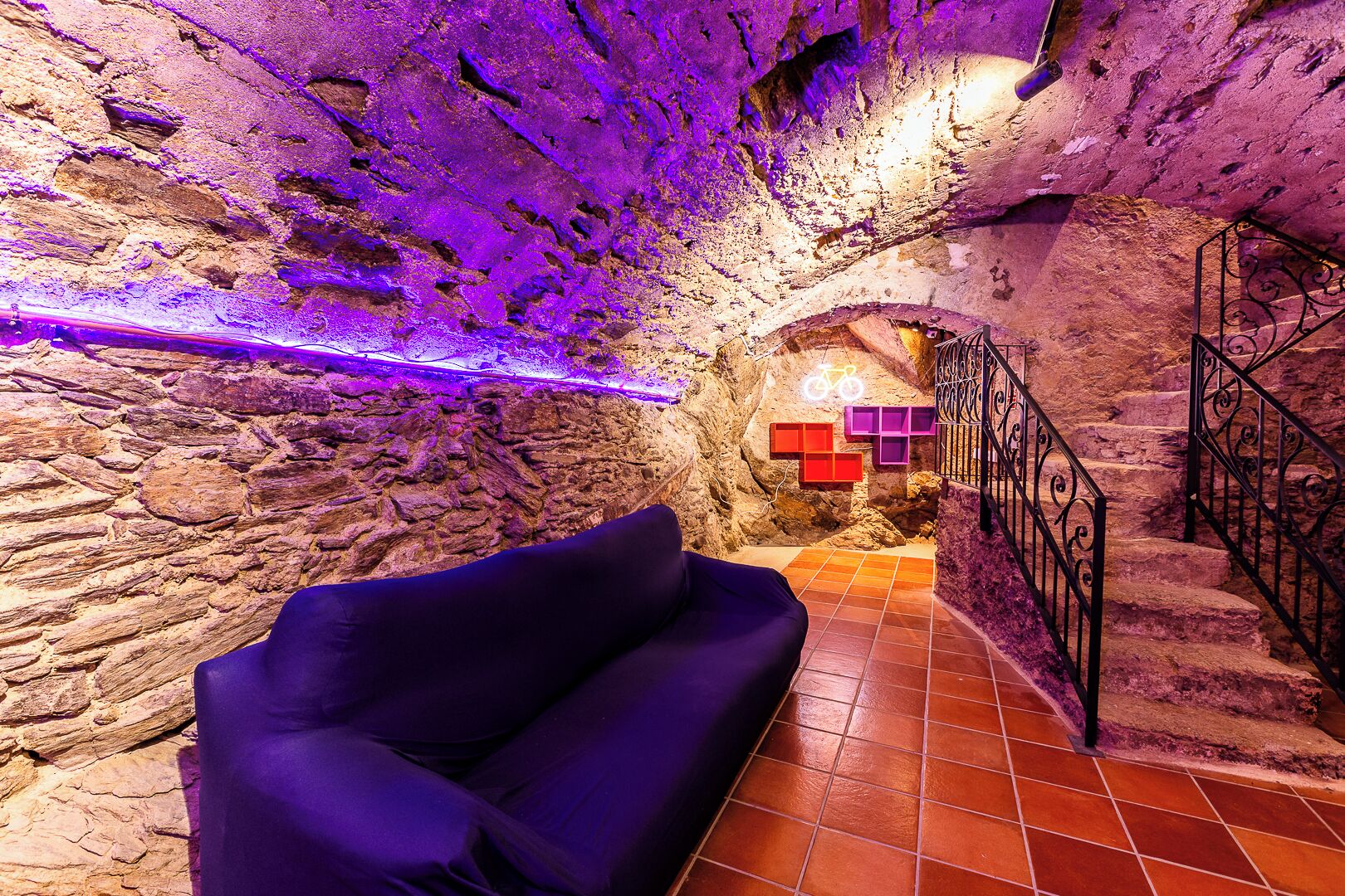 A stone-walled interior with a purple sofa, a staircase, and a neon bicycle sign.