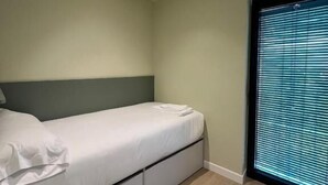 Uma cama de solteiro com cabeceira, uma janela com persianas e um aparelho de iluminação no teto.