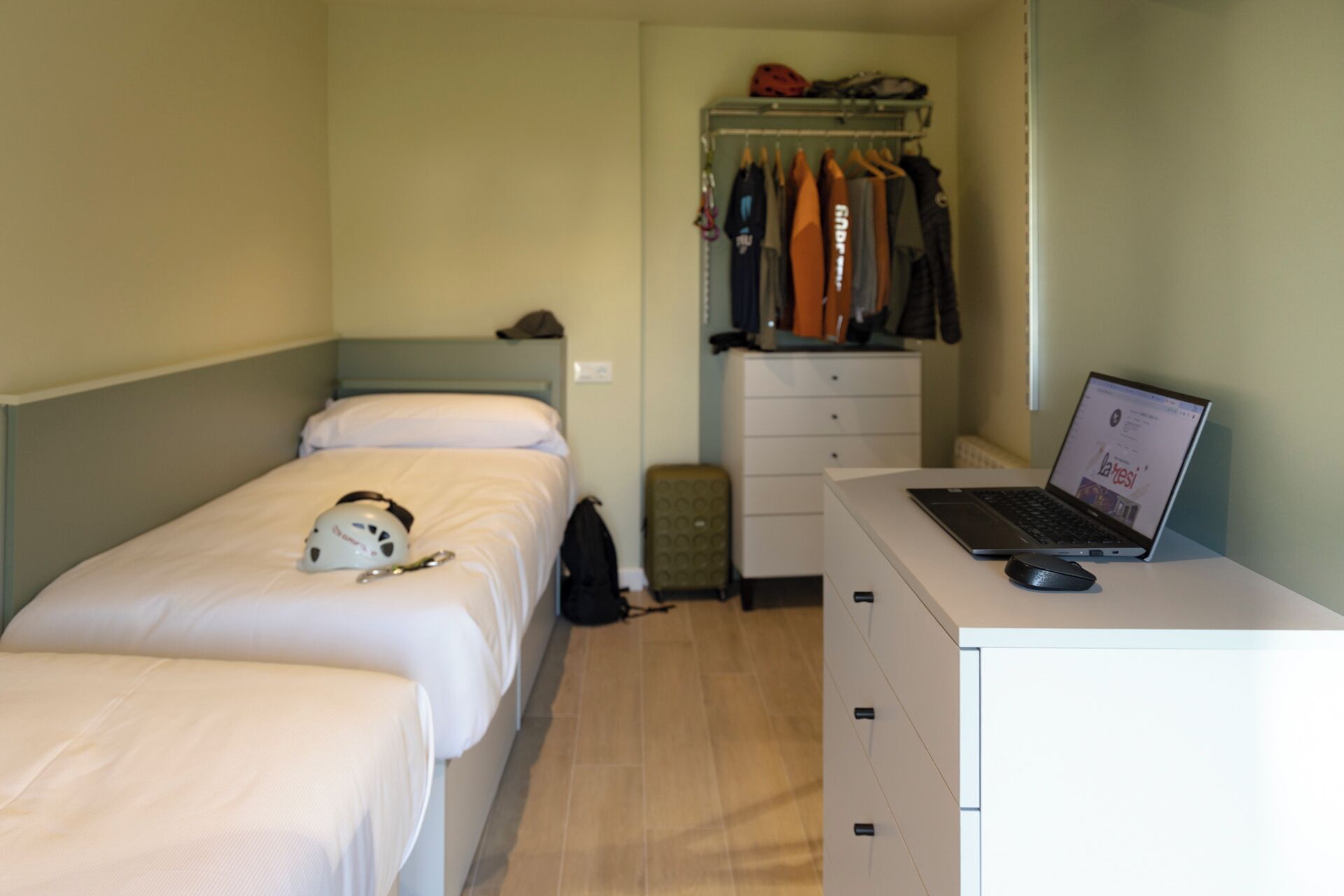 Quarto de hotel pequeno com uma cama, uma escrivaninha com um laptop, uma mala e um cabideiro com casacos.