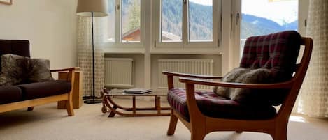 Een woonkamer met een bank, een houten stoel, een tafel en een vloerlamp. Grote ramen bieden uitzicht op de omgeving.