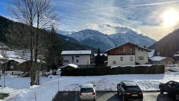 Un parking dont le sol est recouvert de neige, un bâtiment à la façade en bois et, au loin, des montagnes dont les sommets sont enneigés.