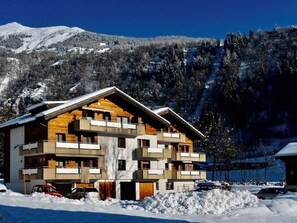 Un hôtel à plusieurs étages doté de balcons, avec une piste de ski en arrière-plan et un paysage montagneux.