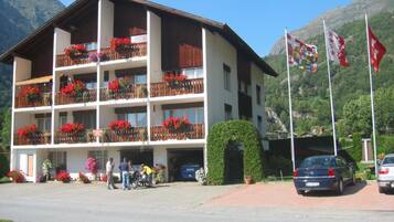 Un hôtel à plusieurs étages, doté de balcons ornés de fleurs rouges, d’un parking et de drapeaux.