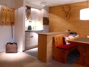 Eine kompakte Küche mit modernen Geräten, eine Wand im Holzdesign mit einem aufgehängten Hirschgeweih und eine kleine Sitzecke mit rotem Kissen.