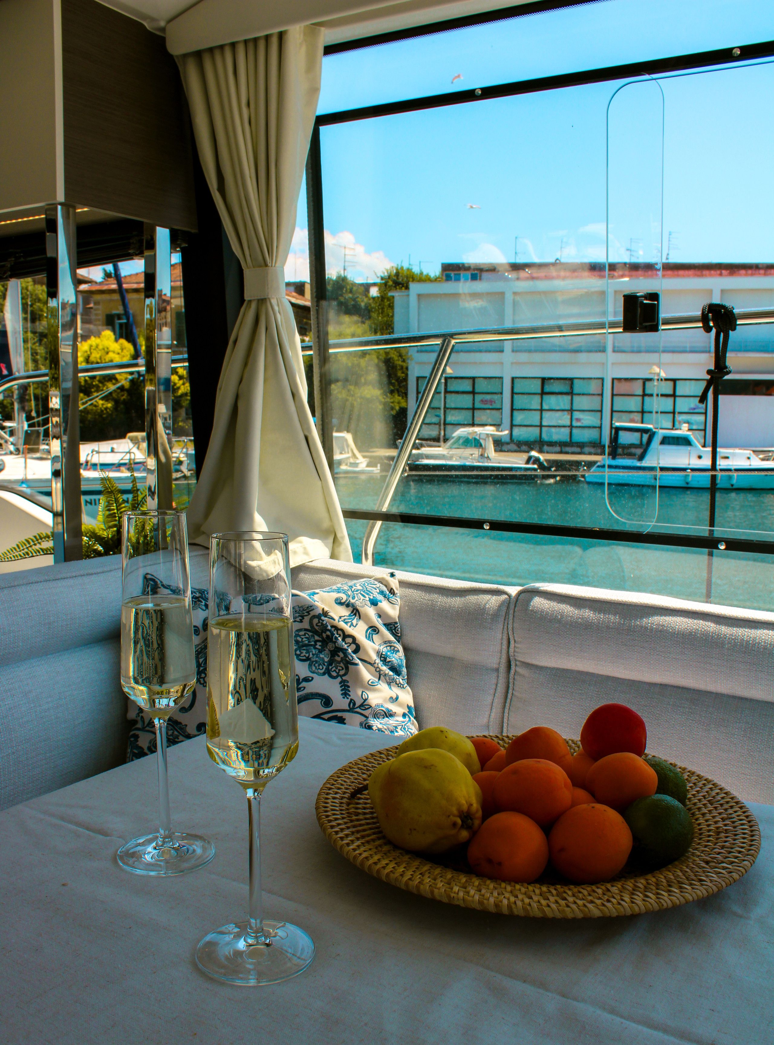A table with two glasses of white wine, a basket of fruit, and a view of a marina with boats and buildings.
