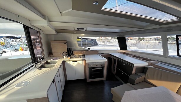 Una cocina pequeña y funcional con pileta, hornilla y alacenas, un espacio de asiento acolchado y vistas a la marina a través de grandes ventanales.