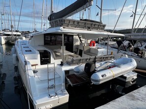 Eine weiße Yacht liegt in einer Marina vor Anker, mit überdachter Sitzgelegenheit, einem kleinen Schlauchboot und einem Rettungsring.