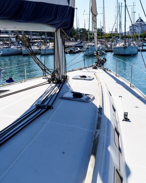Eine Segelyacht liegt in einer Marina an, mit mehreren Booten, wolkenlosem Himmel und einem Gebäude im Hintergrund.
