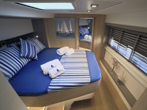 A compact, well-lit cabin with a bed featuring striped bedding and matching pillows, a small window with blinds, and a painting of sailboats on the wall.