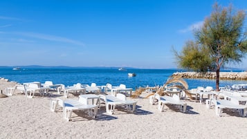 Ein Strand mit weißen Kunststoffliegen, die in Reihen angeordnet sind, eine Palme und eine felsige Küstenlinie im Hintergrund mit Booten.