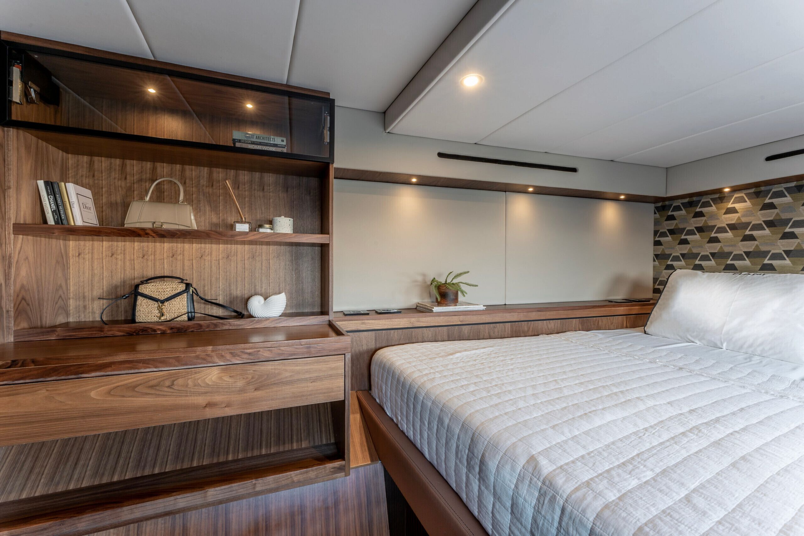 Un dormitorio con una cama grande, estanterías de madera con libros y un bolso, una mesita de noche con una planta, y una pared con estampado geométrico.