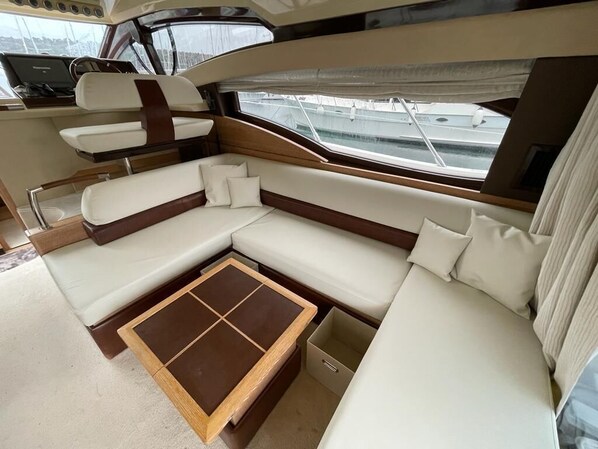 Interior de un yate con un amplio espacio de asientos acolchados, una pequeña mesa y vistas al puerto deportivo a través de las ventanas.
