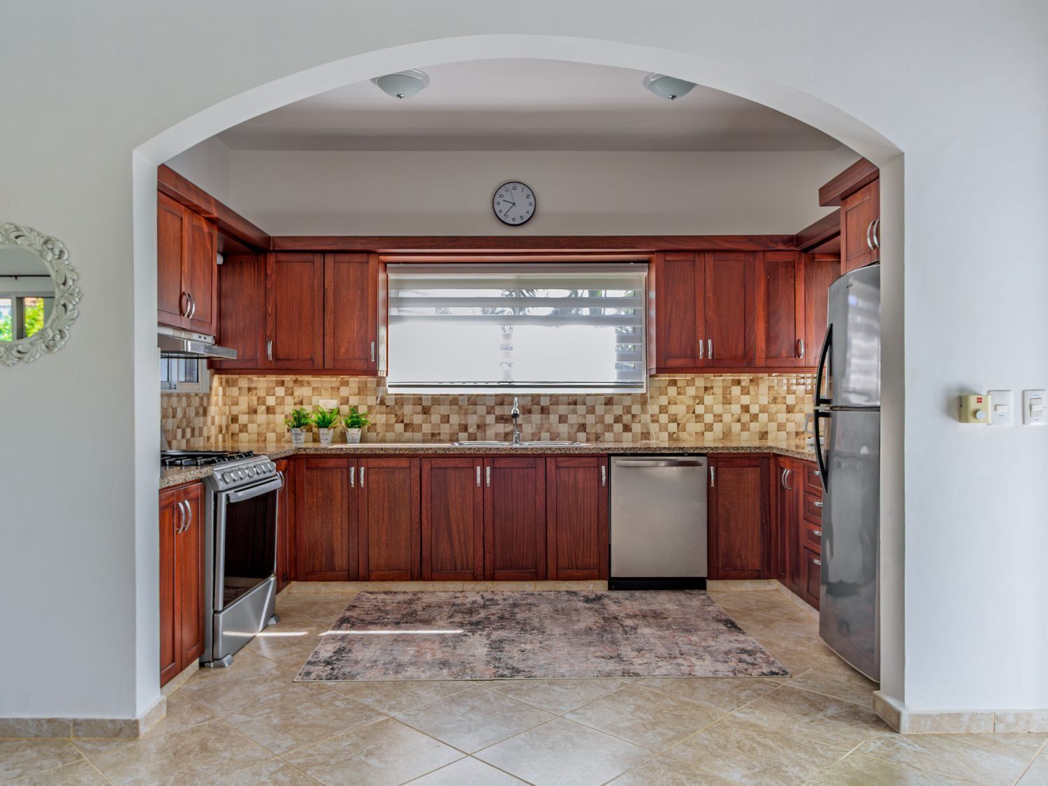 Una cocina con armarios de madera, un refrigerador de acero inoxidable, un lavavajillas y una ventana con persianas.