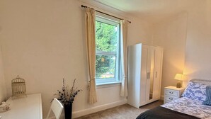Un dormitorio con cama, escritorio, silla, armario, mesita de noche, lámpara y ventana con cortinas.