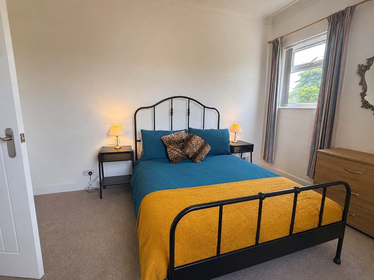 Une chambre à coucher avec un lit, des tables de chevet, une commode, une fenêtre avec des rideaux et une porte.