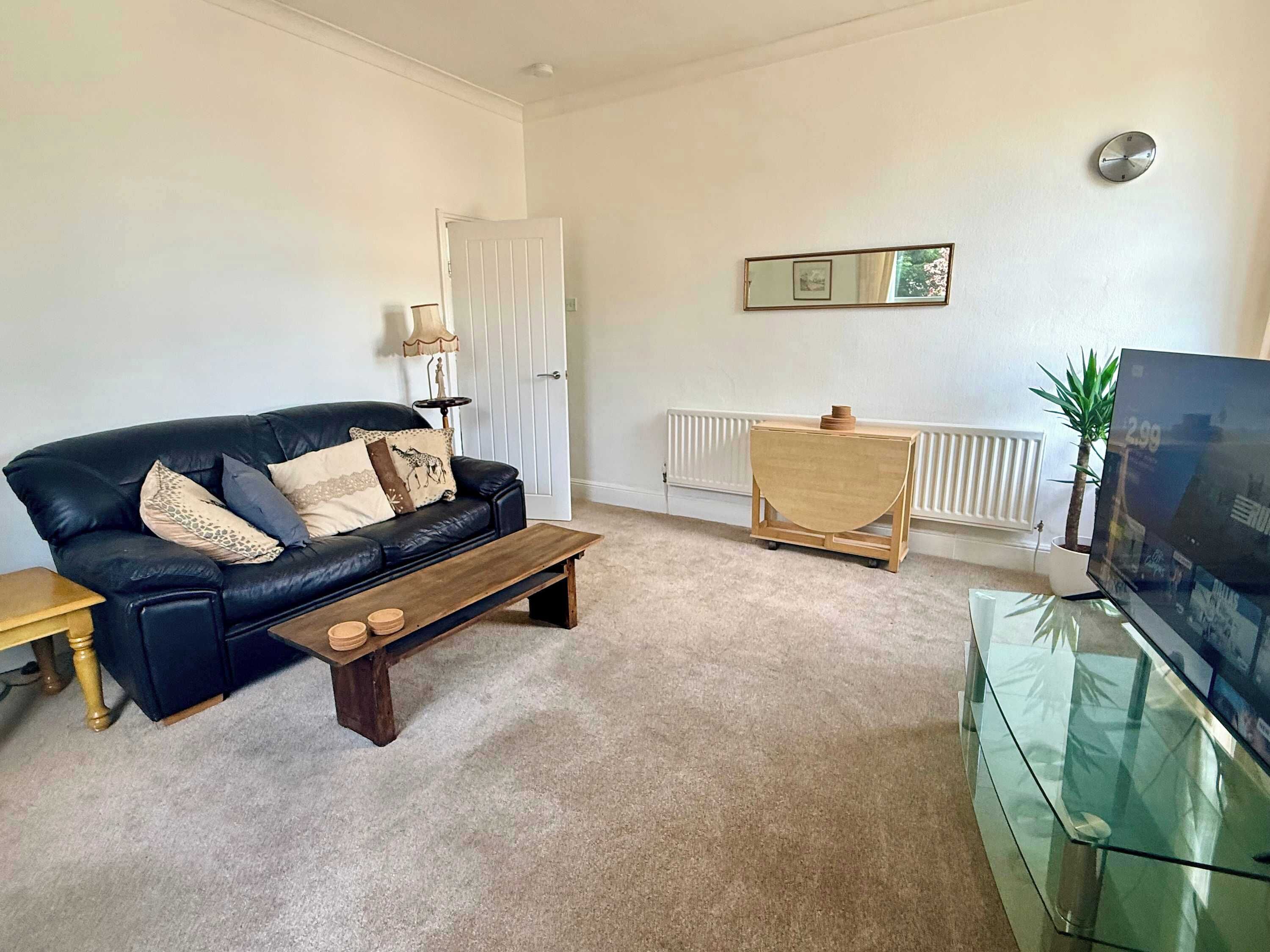 A living room with a black leather sofa, a wooden coffee table, a glass TV stand, a wooden side table, and a radiator.