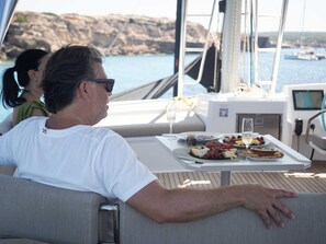 Ein Mann sitzt auf dem Deck eines Bootes, ein Tisch ist für eine Mahlzeit gedeckt, mit Blick auf eine felsige Küste.