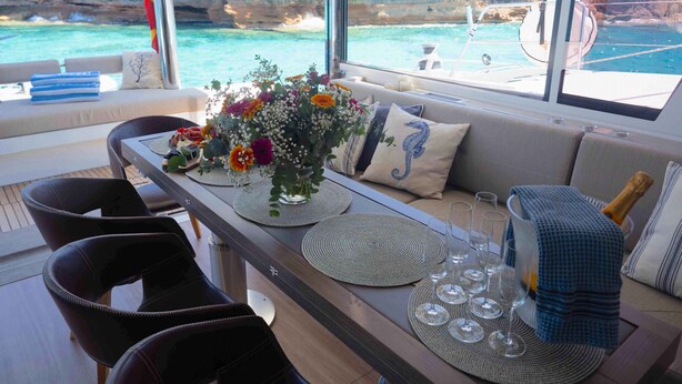 A dining area with a table set for six, a sofa, and a view of the sea and cliffs. - Sun-Kissed Sailing Adventure in Islas Baleares, Mallorca (Andratx)