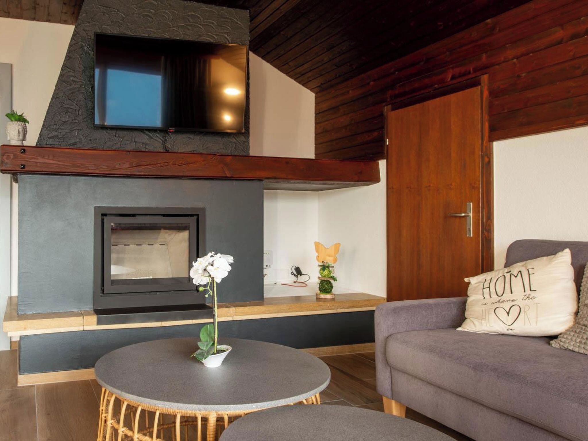 A modern living room with a fireplace, a television mounted on the wall, a round table with a flower, and a sofa with a cushion.