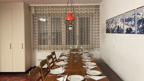 Een eetkamer met een lange houten tafel voor zes personen, omringd door stoelen, met een patroon gordijn en een rode hanglamp erboven.