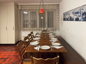 A dining room with a long wooden table set for six, flanked by chairs, with a patterned curtain and a red hanging light fixture above. - Fifinhaus 1. OG Süd Appartement/Fewo, Dusche und Bad, WC, 3 Schlafräume by Interhome (Fiesch)