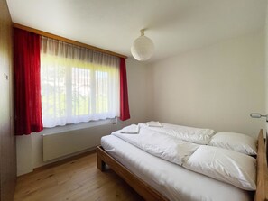 A bedroom with a wooden bed, a radiator, a window with red curtains, and a white ceiling light. - Fifinhaus 1. OG Süd Appartement/Fewo, Dusche und Bad, WC, 3 Schlafräume by Interhome (Fiesch)